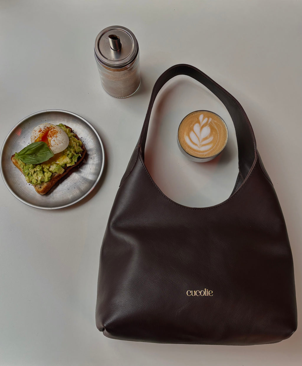 Bolso Jolie Shopper Mini de piel sobre una mesa, junto a un café — Cucolie