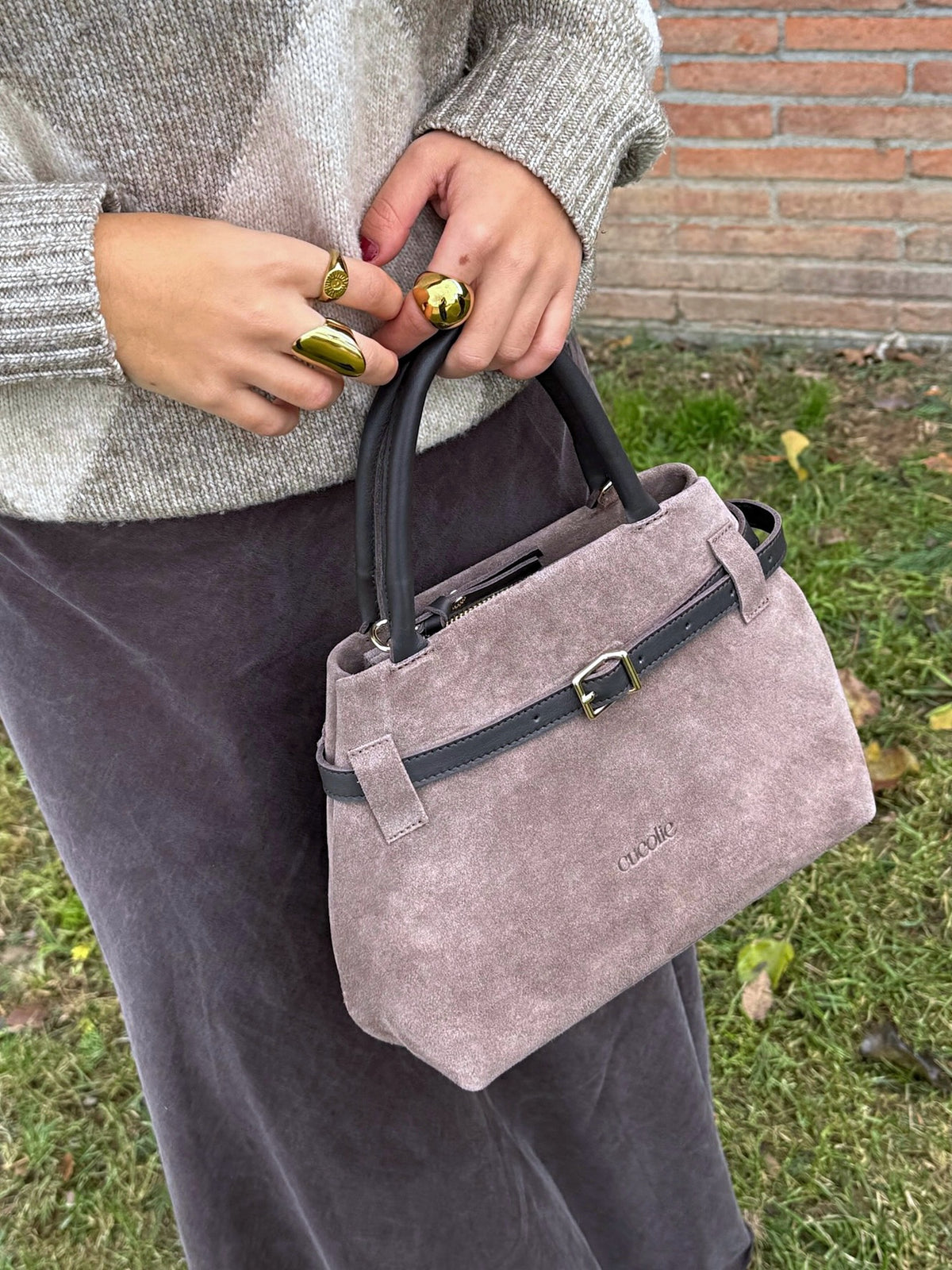Cuco mini de serraje taupe en la mano, junto a una pared de ladrillo