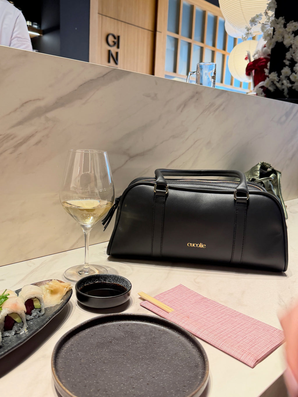 Bolso Cucolie de piel negra sobre la barra de un restaurante, junto a una copa de vino