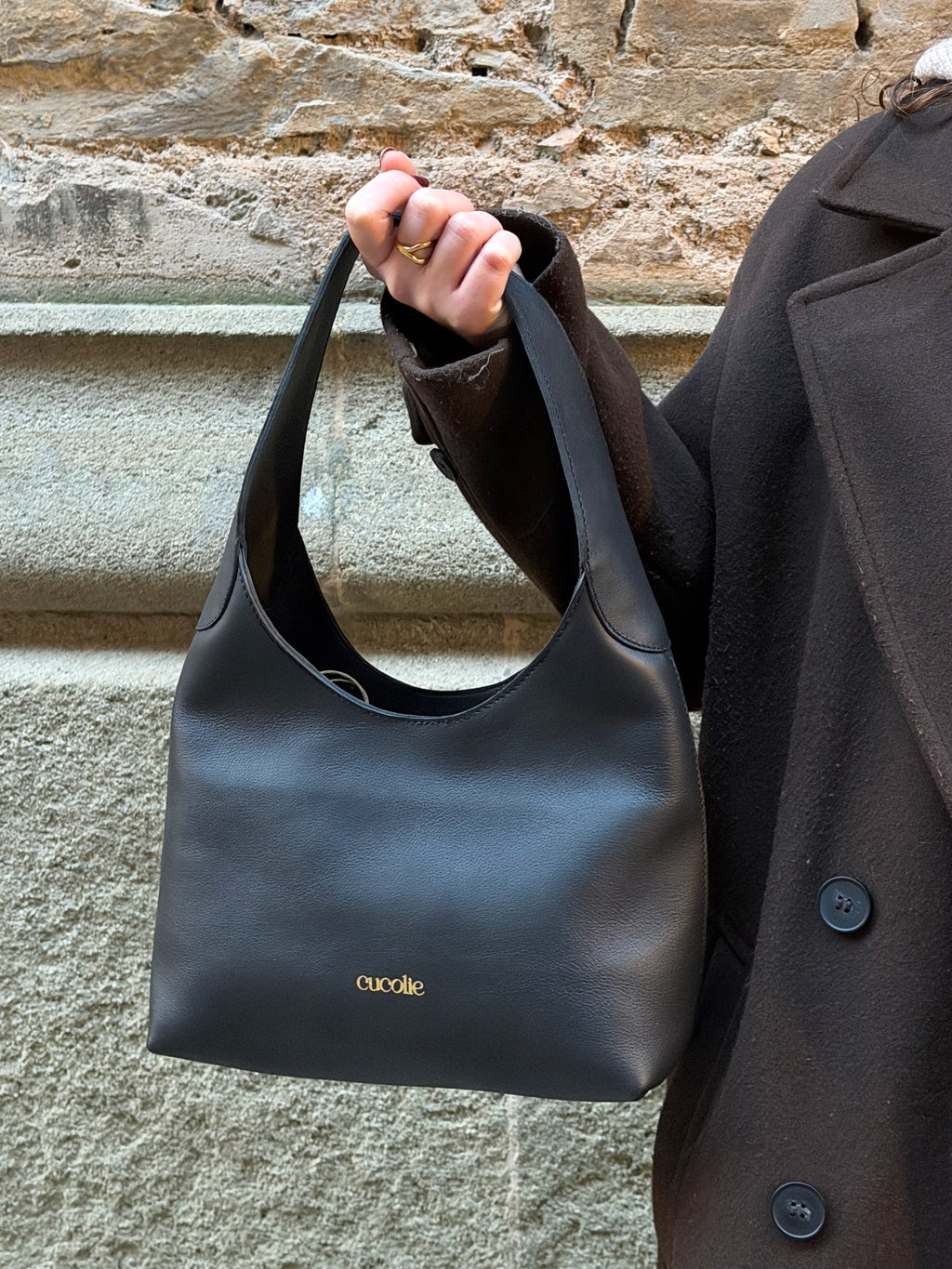 Bolso Cucolie de piel negra sujeto en la mano, frente a un muro de piedra