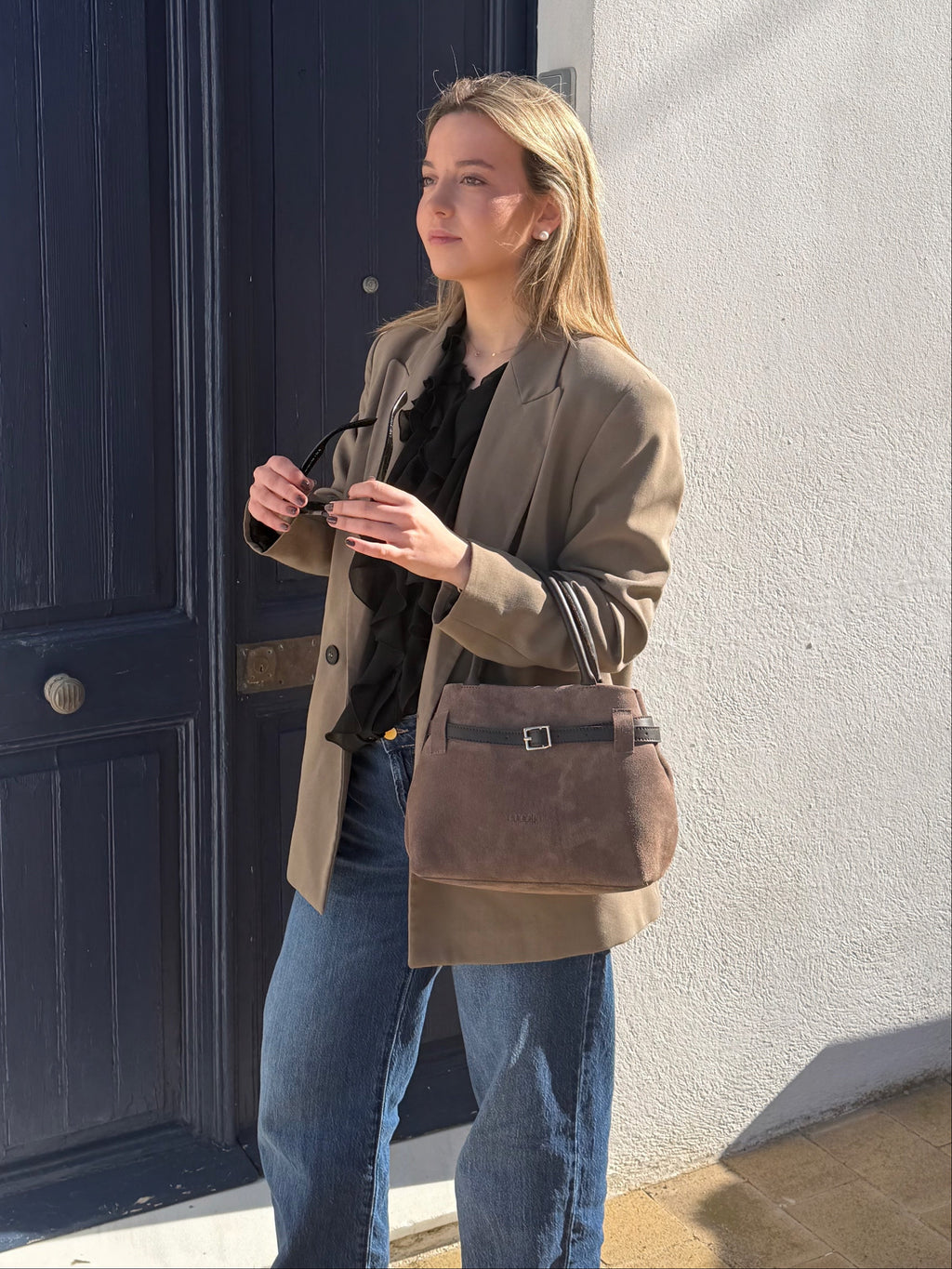 Bolso Cucolie de serraje marrón con americana camel, frente a una puerta azul